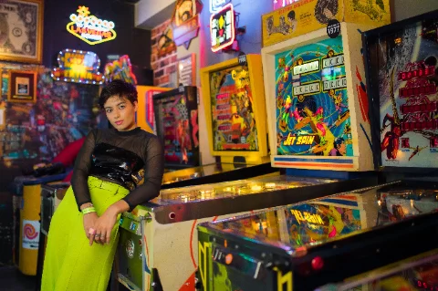 A teenager leaning on a row of lit-up pinball machines in a retro arcade.